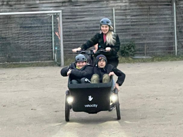 Drei Kinder fahren in einem Lastenrad auf einem Platz.