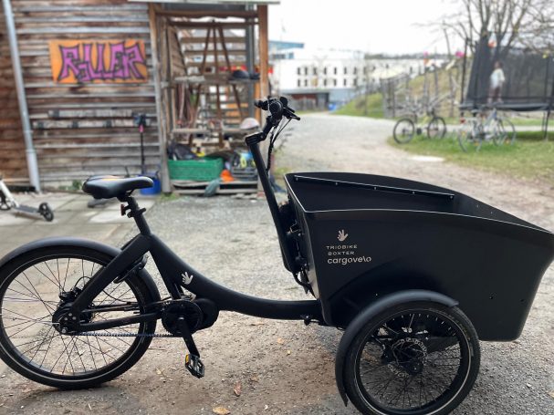 Dreirädriges Lastenrad mit schwarzer Box auf einer schmalen, asphaltierten Straße.